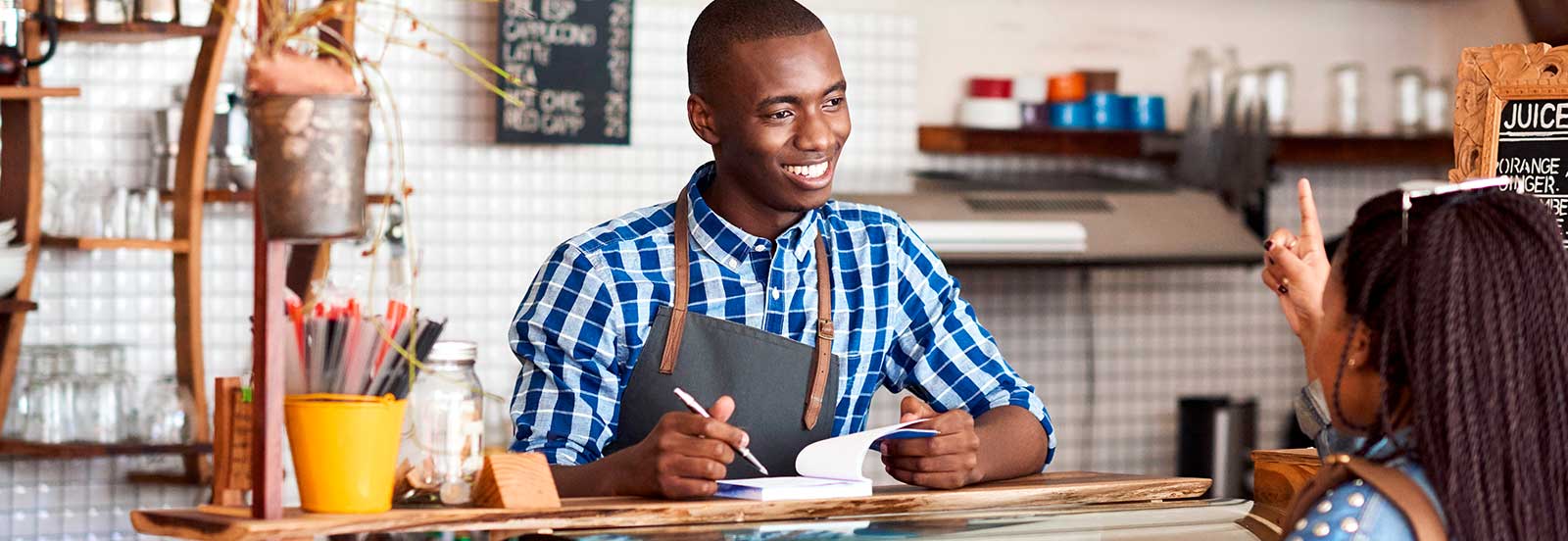 A male barista at the counter in a coffee shop taking an order from a woman. -1600x552.jpg